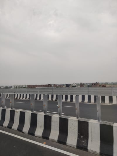 The image shows a highway in India on an overcast day. The foreground features a concrete barrier with alternating black and white diagonal stripes. Vertical grey poles, possibly for lighting or signage, are intermittently placed along the barrier. The road surface is dark asphalt with a white lane marker. In the background, beyond another grey metal barrier, there is a line of buildings, some appearing to be under construction, and some residential structures. The sky is uniformly grey and cloudy.