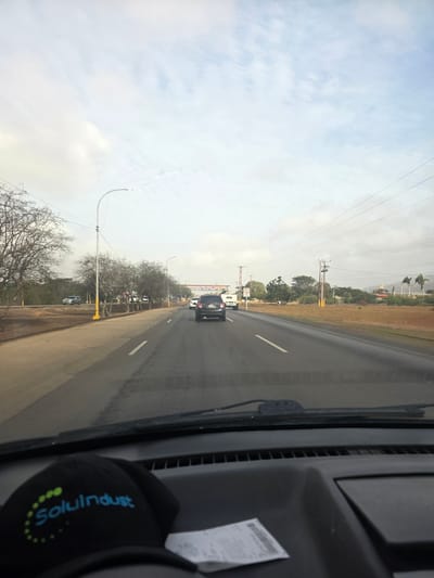 A view from inside a vehicle driving along a multi-lane road. A dark-colored car is visible ahead on the road. The roadside is marked with trees, street lights, and utility poles. The sky is overcast. A black cap with the word "SoluIndust" is present in the foreground. I cannot confirm the location as San Antonio, Venezuela.