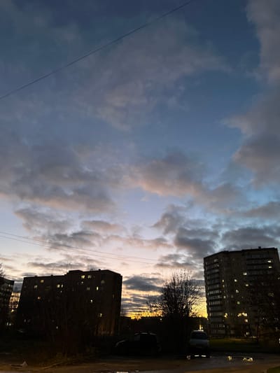 This image depicts an urban residential scene in Voskresensk, Russia, at dusk or early night. The sky transitions from deep indigo at the zenith to pale blue, pinkish-orange, and yellow near the western horizon, indicative of twilight, with extensive stratocumulus cloud cover. A single, dark utility wire is visible diagonally in the upper left sky. The foreground and midground feature two multi-story residential block buildings, silhouetted against the fading light. The building on the left is approximately 7 stories high, and the one on the right is taller, around 10-12 stories, with both exhibiting numerous illuminated windows emitting warm yellow light. Deciduous trees and dense shrubs, largely defoliated, are present between and around the buildings. The ground in the immediate foreground is uneven and wet, with several reflective puddles. Two vehicles are partially visible: a dark-colored sedan or hatchback parked on the left and a light-colored van on the right. No individuals or active interactions are observed within the frame.