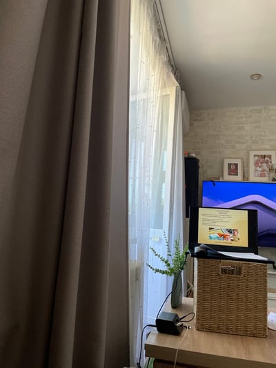 The image shows an interior scene, likely a living room or home office. On the left, a thick, dark brown curtain is drawn. To the right of the curtain, a sheer white curtain is visible, covering a window. Beyond the sheer curtain, a white wall with subtle brick-like texturing is visible, and a white air conditioning unit is mounted high on the wall.
In the foreground, a wooden desk or table is present. On this table, a black laptop is open, displaying a presentation slide with Russian text and an image of a child. To the right of the laptop is a woven wicker basket. A black charging block with multiple cables plugged into it is also on the table, near a small green vase containing a plant with delicate, elongated leaves.
In the background, a large television screen is visible. On top of the TV stand, there are two framed pictures, one of a child and another of two individuals holding a baby. A black speaker or cabinet is positioned to the left of the TV. A digital clock displaying "11:00" is visible on top of this unit. The overall lighting suggests daytime, with light filtering in from the window.
The image does not contain any identifiable landmarks or information to suggest the city of Osinniki, Russia, or any other specific geographical location. The scene appears to be a domestic indoor environment.