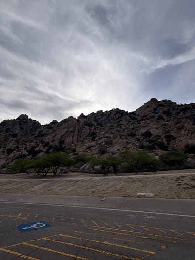 The image shows a cloudy sky over a rocky mountainside with scattered trees. In the foreground, there is a paved road with yellow markings and a blue handicap symbol. The scene appears to be in an outdoor, natural setting, possibly a park or rest area, given the road markings. There are no people visible in the image. The environment is dry and arid, typical of a desert or semi-arid region, with the rocks and sparse vegetation suggesting this. The lighting suggests it is daytime, but the overcast sky creates a subdued atmosphere. The location is specified as Dragoon, United States.