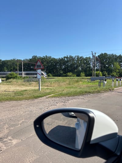The image captures a view from a car's side mirror, showing a rural scene with a railway crossing in Pereiaslavske, Ukraine. A bright blue sky dominates the upper portion of the image. In the middle ground, a train is passing on the tracks, moving from left to right. A red and white triangular warning sign with a depiction of railway tracks is prominently displayed on a post near the crossing. Another white post with red diagonal stripes stands next to it. Trees form a dense line behind the railway tracks. The foreground shows a dirt and gravel road. The side mirror reflects the interior of the car, including a person wearing a light-colored garment. The overall environment appears dry and sunny.