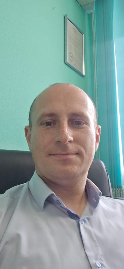 This is a media file featuring a close-up, sideways portrait of a man wearing a light blue collared shirt. He has a shaved head and appears to be in his late 30s or early 40s. A thin, vertical scar is visible on his left cheek. The man is looking directly at the camera with a neutral expression.
The background suggests an indoor setting. The wall behind him is painted a light teal color. A framed document, possibly a certificate or diploma with a circular seal and text, is mounted on the wall to his right. The lighting appears to be artificial, possibly from an overhead source, casting soft shadows. The overall impression is that of a casual selfie taken in an office or personal space. No specific time of day or weather conditions are discernible. The location context of Bryansk, Russia, does not provide any specific visual cues within the image itself.