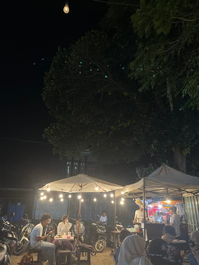 The image depicts a nighttime scene at an outdoor food stall located in Tambonbaroh, Indonesia. The stall is illuminated by a string of lights draped over a white umbrella, casting a warm glow. Several people are visible, some seated at tables and others working at the stall. Motorcycles are parked in the background, suggesting a street-side location. A large tree with visible fruit, possibly jackfruit, looms over the scene. The overall atmosphere appears casual and social, typical of a local gathering spot.