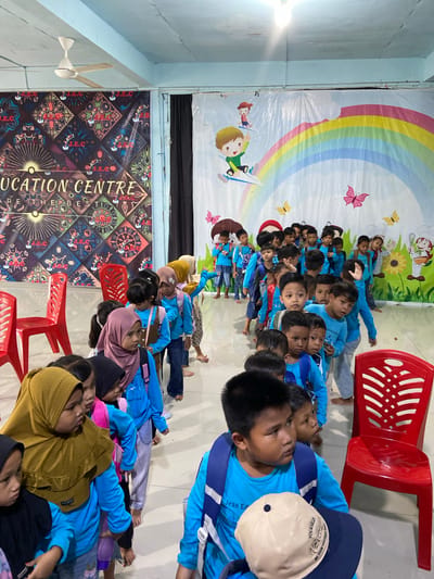 In Tanjung Gading, Indonesia, children in blue shirts line up in a hallway with a colorful mural depicting a rainbow and cartoon characters on the wall. A banner for "EDUCATION CENTRE" is visible to the left. Red plastic chairs are placed along the right side of the hallway. The children, wearing backpacks, appear to be waiting or moving in a procession. The environment seems to be an indoor space, possibly a school or community center, illuminated by overhead lighting.