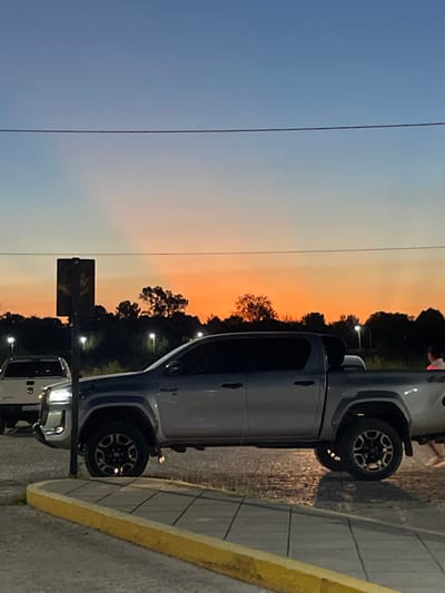 A silver Toyota Hilux truck is parked on a gray paved surface with a yellow curb. The sky transitions from blue to orange in the background, indicative of sunset. A power line stretches across the upper-left quadrant of the image. Several trees and a street lamp are visible in the background, alongside another silver vehicle.