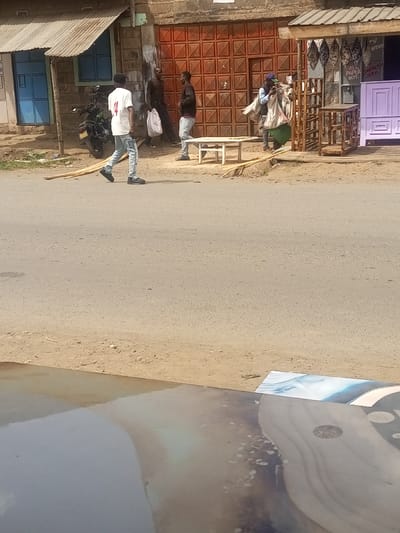 The image depicts an outdoor street scene within Kiamaina ward, Kenya, under bright daylight conditions. In the foreground, a paved asphalt road extends across the frame, with a dirt shoulder on both sides. The bottom portion of the image shows a blurred, reflective surface, likely a tabletop or dashboard from which the photograph was captured.

In the midground, several individuals and structures are visible along the dirt shoulder. From left to right:
*   A male subject, wearing a white t-shirt with a number "4" on the back, light blue ripped jeans, and dark shoes, walks from right to left across the paved road.
*   Behind him, parked on the dirt shoulder in front of a building with a corrugated metal roof and blue window/door, is a black motorcycle.
*   Adjacent to the motorcycle, in front of a building with a large, reddish-brown patterned metal gate, two male subjects are standing. One, wearing a dark t-shirt and trousers, holds a white bag in his left hand. The other, wearing a dark long-sleeved shirt and light blue jeans, faces him.
*   To their right, on the dirt shoulder, a small white wooden table with simple legs is present.
*   Further right, a male subject wearing a blue cap, light-colored long-sleeved shirt, and a green garment (possibly a skirt or kikoi) is carrying multiple large, light-colored sacks over his shoulder, moving towards the right side of the frame.
*   He is passing in front of a shop with a corrugated metal overhang. The shop displays various items including wooden shelving units, a purple headboard or bed frame, and printed papers/posters on the wall, some advertising "PAPER COMPRESS SERV" and "TV". Decorative shields are mounted above the shop's display area.
*   Several long, thin wooden poles or beams lie horizontally on the dirt shoulder in front of the white table.