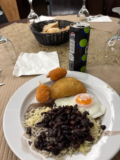 A dining scene is depicted on a table in Santa Maria da Feira, Portugal. The table is covered with a patterned brown tablecloth. A white, circular plate holds a meal consisting of two breaded and fried shrimp with tails, one golden-brown fried pastry or rissole, and a sunny-side-up fried egg. Below these items, a bed of rice is mixed with black beans and their accompanying dark sauce. Adjacent to the plate, a crumpled white paper napkin lies flat. Further back on the table, a black woven basket contains several pieces of bread. An open 330ml black Pepsi can with green and blue polka dots stands upright. Multiple clear wine glasses are visible, some inverted, and a fork is partially visible on the left. The background is blurred, showing dark wooden furniture or wall elements. No individuals are present in the frame.