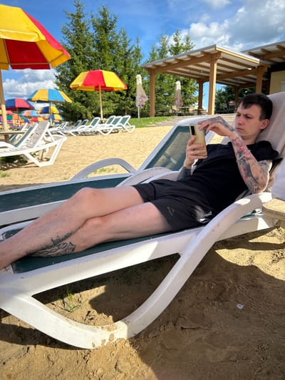 A young man with multiple tattoos on his arms and legs is relaxing on a white lounge chair on a sandy beach. He is wearing a black t-shirt and black shorts and is holding a smartphone in his hands, looking down at it. The background shows several beach umbrellas in red, yellow, and blue, and a wooden structure with a covered roof. Trees are visible behind the structure. The scene appears to be a recreational area, possibly a beach resort or park in the city of Yubileyny, Russia. The lighting suggests it is a sunny day with some clouds in the sky.