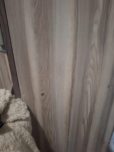 This media file is a close-up, medium-angle shot of a light-colored wood-grain surface, likely a shelf or countertop. The wood grain features a pattern of light brown and grey wavy lines, with some darker streaks interspersed. There are at least three visible screws securing the surface, one towards the top left, another in the middle right, and a third further down on the right. The surface appears to be part of a piece of furniture or cabinetry. In the lower right corner, a fuzzy, cream-colored textile is partially visible, possibly a blanket or towel. The lighting suggests an indoor setting, and the shadow cast on the wood surface indicates a light source from the upper right. There is no visible text in the image. The context of Ötegen batyr, Kazakhstan, does not provide specific visual cues within the image itself.