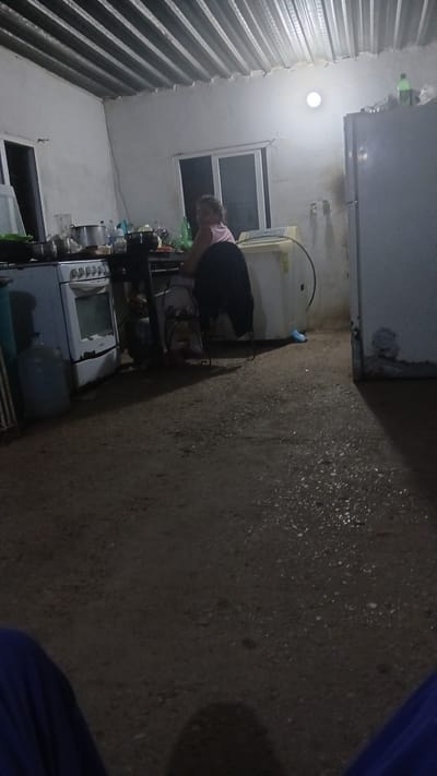 The image depicts a room in what appears to be a residential setting, potentially in the city of Putucual, Venezuela, though this cannot be verified with certainty from the image alone. A woman is visible, bending over in the vicinity of a stove and other kitchen appliances. The kitchen appears cluttered, with various items on shelves and surfaces. The walls are painted white, and the ceiling is constructed of corrugated metal. The lighting is poor, and the ground is mostly out of view.