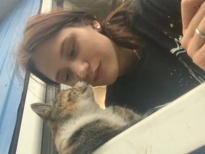 In this close-up, low-angle shot, a young woman leans down to nuzzle her cat. The woman has light brown hair pulled back in a ponytail and is wearing a black t-shirt with colorful speckles. She has a small gold earring in her left ear and a silver ring on her right ring finger. Her eyes are closed, and her lips are slightly parted as she connects with her pet.
The cat, with its distinctive tabby markings of brown, grey, and white, is positioned slightly below and to the left of the woman's face. Its eyes are also closed, and its nose is touching the woman's nose, indicating an affectionate moment. The cat's fur appears soft and fluffy.
The background is blurred, but hints of the setting can be discerned. To the left, there is a blue, corrugated surface, possibly an outdoor wall or structure. To the right, a light-colored, possibly white, ledge or surface is visible, upon which the cat seems to be resting. The lighting suggests it is daytime, likely outdoors, with natural light illuminating the subjects. The overall mood of the image is tender and intimate, capturing a shared moment of affection between the woman and her cat. No text is visible in the image.