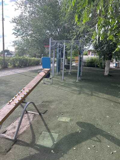 This is an outdoor image of a playground in Saran, Kazakhstan. In the foreground, a wooden seesaw with yellow grips on metal strips is tilted. The playground surface is a green, rubberized material with some worn patches and scattered leaves. Behind the seesaw, a metal structure with monkey bars and a basketball hoop is visible. Trees with green foliage surround the playground. In the background, residential buildings and a paved walkway can be seen. The sky is partly cloudy.