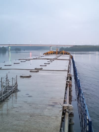 The image depicts a barge on a river, possibly at dusk or dawn, with a bridge in the background. The barge appears to be loaded with concrete slabs, some of which are covered. The riverbanks are lined with trees, and there are lights visible on the bridge. The sky is overcast, creating a muted color palette. The water is calm, with ripples indicating movement. The scene appears to be located in Ivanov Bor, Russia, given the context provided. There are no people visible in the image.
