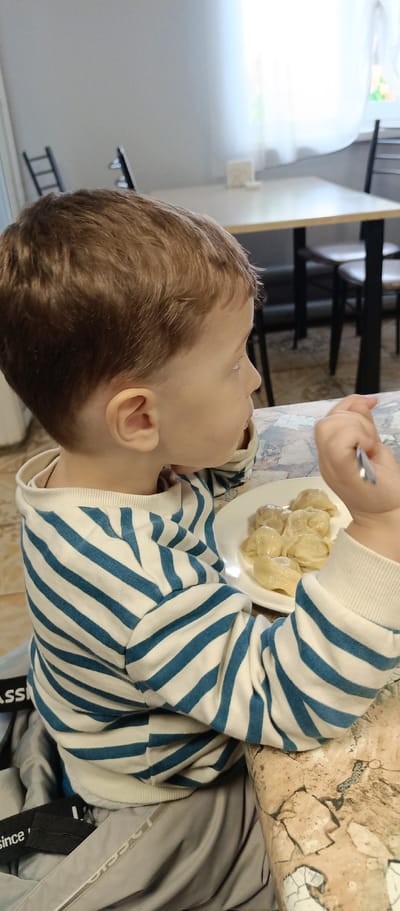 A young child with brown hair and wearing a blue and white striped long-sleeved shirt sits at a table, presumably in a restaurant or cafeteria, and eats dumplings (pelmeni) with a fork. The plate of dumplings is in front of the child on a marbled-patterned table. There are tables and chairs in the background, and a window is visible. The location is in the city of Irbit, Russia.