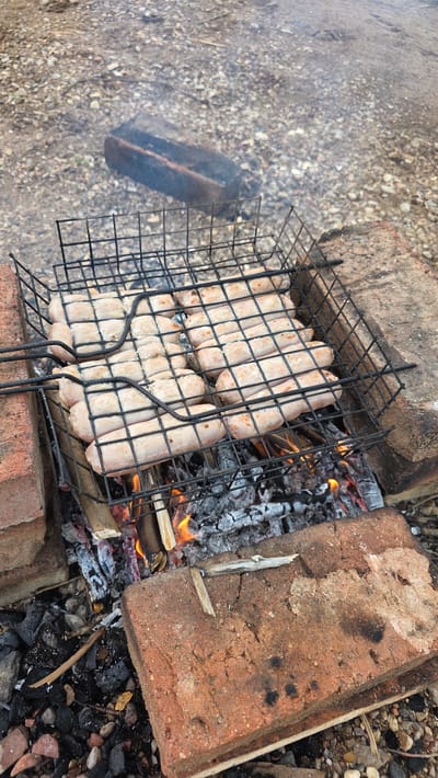 A close-up, high-angle shot shows sausages cooking on a barbecue grill over a fire in what appears to be an outdoor, possibly rural or semi-rural setting, given the gravel and dirt ground. The grill is a black wire rack, holding at least twelve uncooked sausages arranged in rows. The sausages are pale pink and plump. The grill rests on a makeshift structure of red bricks and burning wood, with visible coals and flames underneath. Smoke rises from the barbecue. In the background, a dark, rectangular object, possibly a brick or piece of wood, lies on the gravelly ground. The overall scene depicts food preparation outdoors. The location is specified as Дарьино, Russia.