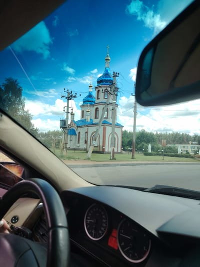 An exterior, eye-level, medium shot taken from the interior of a vehicle shows the Church of the Intercession in Kapitanivka, Ukraine. The church is a light-colored brick building with a white facade and red trim, featuring multiple blue domes topped with gold crosses. The sky is a bright blue with scattered white clouds. In the foreground, the car's steering wheel, dashboard, and side-view mirror are visible. There are utility poles and wires in front of the church. The church is situated on a grassy area with trees in the background. The overall scene depicts a sunny day with the church as the central focus.