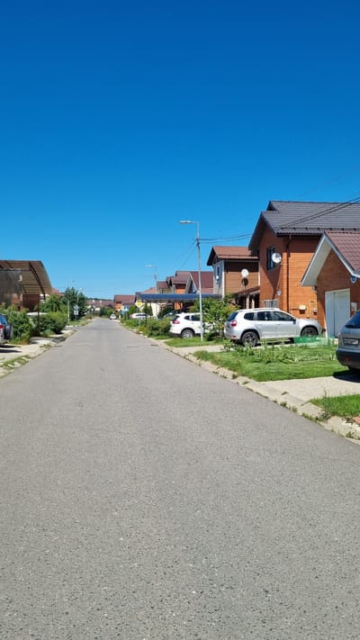 The image depicts a quiet residential street in the village of Verzilovo, Russia, on a clear, sunny day. The asphalt road runs towards the horizon, flanked by well-maintained houses with terracotta-tiled roofs. Several cars are parked along the street and on the lawns in front of the houses. These include a silver SUV, a white SUV, and the rear of a silver car. Lush green trees and bushes line the street, interspersed with streetlights and power lines. The sky is a vibrant, cloudless blue. The scene conveys a sense of peaceful suburban life.