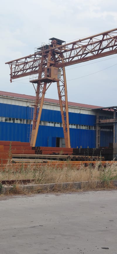 An outdoor industrial scene in Ohangaron, Uzbekistan features a large, rusty orange gantry crane positioned in front of a blue and white industrial building. The building has a row of windows and a red roof. In the foreground, stacks of metallic pipes and beams are piled on the ground, with dry grass and weeds growing in front of them. The ground is a mixture of asphalt and dirt. The sky is overcast.