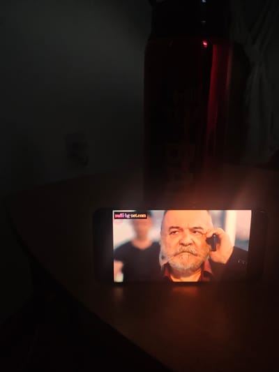 A horizontal smartphone, displaying a close-up of a Caucasian man with a gray beard and mustache, is placed on a table. The screen also displays the URL "nufi-bg-net.com" in the top left corner. Behind the phone is a dark, cylindrical object, possibly a water bottle. The room is dimly lit, with a wall visible in the background. The specific location in Swindon, United Kingdom is not discernible from the image.