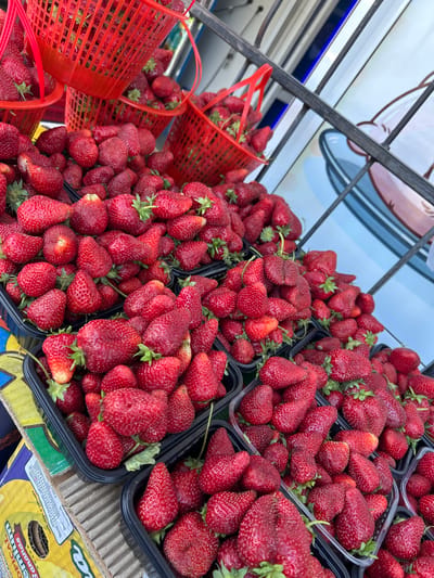 The image shows a large quantity of fresh strawberries displayed for sale in an outdoor market. The strawberries are arranged in black plastic containers and orange plastic baskets. The containers and baskets are overflowing with the red, ripe fruit. The background includes a metal structure and a box with writing in a foreign language (possibly Russian) and the word "Bananas" visible. The exact location is unclear; a more detailed view of the background would be required to determine if the location is in the city of Aqsu, Kazakhstan.