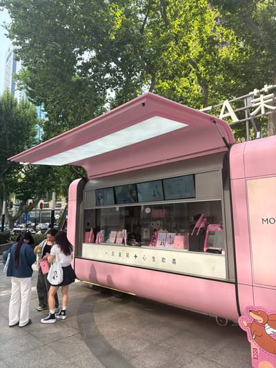 Here is a detailed description of the image:
**Overall Scene:**
The image shows a pink pop-up store or kiosk on a city street in the Xuanwu District, China. There are a few people standing near the kiosk, likely customers. Trees and urban architecture are visible in the background.
**Detailed Elements:**
* **Pink Kiosk:** The kiosk is predominantly pink and has a modern, possibly trendy design. It has large windows displaying items inside, including drink menus and merchandise. It looks to be a food and drink establishment, potentially a tea or dessert shop. The name MOLLY TEA is partially visible. The Chinese characters "一见茉莉心生欢喜" which translate to "Love at first sight with jasmine" are visible on the side of the kiosk, along with the word DOE and a cartoon dog.
* **People:** There are three people standing around the kiosk. One is wearing a white shirt, dark skirt, and black shoes with white socks. Another is wearing a navy blue jacket and white pants. The third is between the two people, and is partially obscured. They appear to be in conversation or interaction with the kiosk, potentially ordering or looking at the menu.
* **Environment:** The setting is an urban environment, likely a commercial area or shopping district. Trees are visible, suggesting a sidewalk or park-like setting. The sky is not clearly visible. Modern buildings are faintly visible in the background.
* **Location:** The image is in the Xuanwu District, China.
* **Signage:** A sign with Chinese characters and English letters is partially visible near the top of the frame.
* **Layout:** The angle suggests the photo was taken from a slightly low vantage point. The kiosk is oriented in a way that draws attention to its display windows and the people interacting with it.