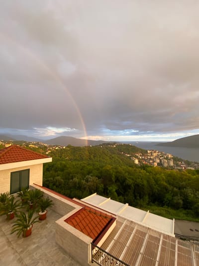 This image captures a breathtaking panoramic view from a high vantage point, likely a terrace or balcony, overlooking a coastal or lake region in Trebesin, Montenegro.
In the immediate foreground on the right, parts of a building with a distinctive red-tiled roof and white facade are visible. A balcony with dark railings extends from this structure, alongside a translucent, corrugated roof, possibly covering a patio or extension. Further towards the center-right foreground, another section of a building features a red-tiled roof, a light-colored wall, and a dark green-shuttered window. A spacious patio area on this level hosts several vibrant potted plants, resembling sago palms, in terracotta pots.
The scene then opens up to a vast natural landscape. Lush green hills, densely covered in trees and vegetation, slope down towards a body of water. A town or settlement, characterized by numerous white and light-colored buildings, is nestled along the shoreline and extends up the lower slopes of the hills. In the far background, more mountains rise from beyond the water, their peaks partially obscured by the sky.
The sky is a prominent feature, displaying a dramatic mix of light and shadow. Dark, stormy clouds dominate the left side, gradually transitioning to brighter, sun-kissed patches towards the right. A magnificent, vivid rainbow arches across the sky, its colors clearly visible, adding a magical element to the landscape.
The lighting suggests late afternoon or early evening, with a soft, golden glow illuminating parts of the clouds and distant hills, indicative of the sun breaking through after recent rain. The presence of the rainbow strongly points to recent rainfall. The overall atmosphere is one of serene natural beauty and wonder. There is no visible text in the image.