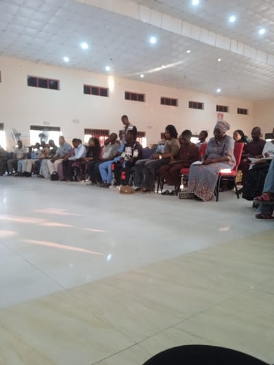 An indoor space with rows of people seated. A diverse group attends, including men, women, and children of various ages. Many sit in chairs, while one stands. Light streams in, casting shadows on the tiled floor. Overhead, numerous lights illuminate the room and rectangular windows punctuate the high walls. The space appears to be a church or auditorium.