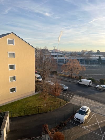 The image presents a panoramic view of an urban landscape, likely in Schweinfurt, Germany, under a bright, clear sky.

In the foreground, a light yellow residential building with a dark tiled roof occupies the lower left. Its facade features several rectangular windows. Adjacent to this building is a paved area, functioning as a parking lot and road, where various vehicles are parked. A prominent white SUV, possibly a BMW, is seen in a parking spot on the right. Further back, a white van and several other grey cars are also visible. A strip of green grass and numerous bare deciduous trees, devoid of leaves, line the paved areas, suggesting late autumn, winter, or early spring. A few small figures, likely people, can be discerned walking on a path or street in the mid-distance.

Dominating the mid-distance on the left is a tall, slender industrial chimney, actively emitting a plume of white smoke or steam that drifts across the sky. Behind the trees and vehicles, a cluster of industrial or commercial buildings with large windows stretches across the middle ground. The background reveals a hazy skyline of distant buildings, indicating the wider city or town.

The scene is captured during daytime, with clear sunlight casting shadows on the paved surfaces and buildings. The sky is a bright blue, adorned with wispy white clouds and several faint contrails. No specific event is taking place beyond the ordinary activities of an urban environment. There is no visible text in the image.