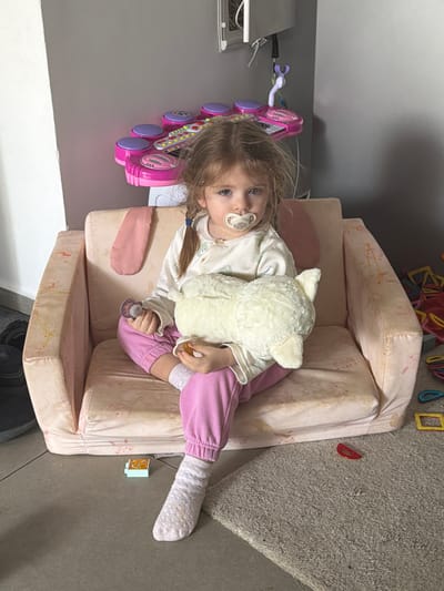 A young child lies on a small, tan-colored couch. She wears a white top, pink pants, and patterned socks. She has a pacifier in her mouth and holds a white stuffed animal. A pink toy guitar sits beside her. The couch is situated on a carpeted floor. In the background, there is a gray wall with a mounted unit. Located in a residence in Harish, Israel.