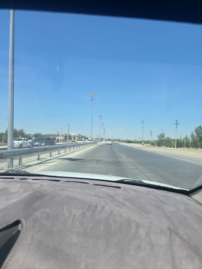 The image captures a view from inside a vehicle, looking out at a road in Ganchkash, Uzbekistan. The scene is dominated by a clear, bright blue sky and a wide asphalt road stretching into the distance. Several vehicles, including cars and a white van, are visible on the road. Along the sides of the road, there are streetlights, some equipped with solar panels, and utility poles with power lines. Vegetation, including trees and some sparse greenery, lines the roadside. In the foreground, the vehicle's dashboard is partially visible, showing its textured surface. The overall impression is of a sunny day in a semi-arid or desert environment within a city or town.