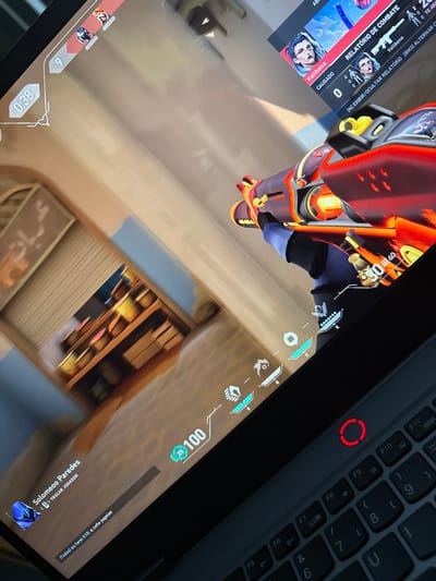 The image shows a laptop screen displaying the video game Valorant. The player character holds a vibrant, fiery weapon. In-game, the player has full health (100) and some credits (25). A timer indicates 0:38 remaining. The player's username is "Solomeoo Paredes," and an option to trade characters exists. The keyboard is visible to the right, with a red-circled power button. Game statistics and character names ("Vallbona") appear on the screen's edges.