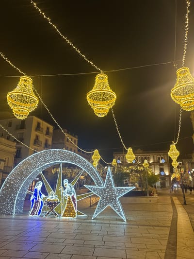 Nighttime scene showcasing holiday decorations on a street. Overhead, golden light fixtures dangle from wires, forming a festive canopy. Sidewalks are made of light-colored stone. A large, white light display on the ground depicts a star, crescent moon, and figures in robes, possibly representing the Nativity. Buildings are visible in the background, some adorned with similar light decorations. The overall impression is one of celebratory illumination.