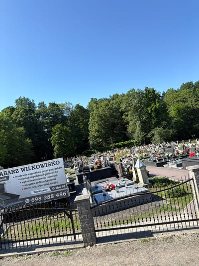 The image shows a cemetery in Wilkowisko, Poland, under a clear blue sky. In the foreground, a white sign with black text displays services offered, including dismantling tombstones, installing tombstones and graves, creating earthly graves, and using all types of stone, inscriptions, and engravings. The sign also shows a phone number: 698 388 486 and the text "Gracie 92 Zapraszamy". Behind the sign, a black wrought-iron fence encloses a section of the cemetery, which is filled with numerous gravestones and floral arrangements. Lush green trees form a dense backdrop to the cemetery. The ground is covered with gravel and some patches of grass. The overall scene is tranquil and solemn, consistent with a place of remembrance.