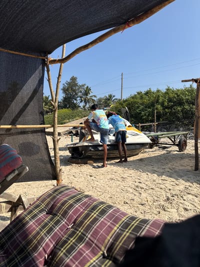 Two men in t-shirts and shorts stand beside a small boat on a trailer. The boat and trailer rest on sandy ground. Overhead, a structure of bamboo and black netting provides shade. A person's body and plaid cushion are in the foreground, partially obstructing the view. Trees are visible in the background, and the sky is clear.