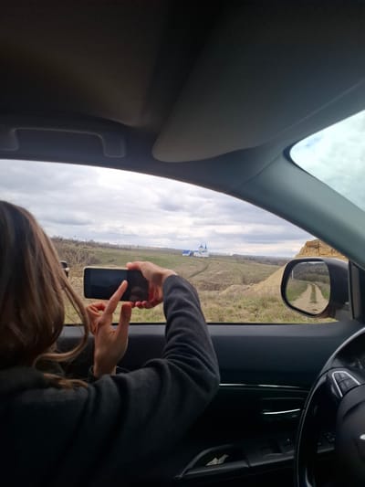 The image captures a scene from inside a vehicle, looking out a side window at a rural landscape. In the foreground, a person, likely a woman, is holding a light-colored smartphone with a dark screen and appears to be taking a photograph or video. Her hands are positioned on the phone, and part of her brown hair and a dark long-sleeved garment are visible.

The car's interior features include a dark A-pillar on the left, a dark door panel on the right, and a partial view of the steering wheel and dashboard in the top right corner. The exterior rearview mirror is also visible, reflecting a dirt road winding through dry, grassy terrain.

Outside the car, the landscape consists of rolling hills covered in sparse, dry vegetation and light-colored grass, characteristic of late autumn or early spring. A dirt path or road is visible winding through the hills. The sky is overcast with a mix of white and grey clouds. In the mid-distance, a prominent church with distinctive blue domes stands out against the horizon, its architectural style indicative of an Orthodox place of worship. The setting appears serene and open, with distant trees marking the horizon.

The activity shows the individual documenting the scenic view, specifically the church. There is no discernible text visible in the image. The weather is cloudy daytime. The location context indicates this scene is in or around Yablonovo, Russia.
