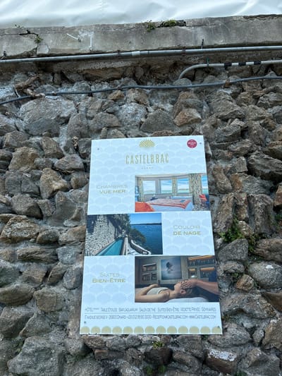 The image shows a sign for the Hotel Castelbrac, a hotel located at 7 avenue George V, 35800 Dinard, France. The sign is attached to a stone wall. The sign is white and features a blue and white pattern. It has the name of the hotel at the top in bold letters, followed by a small image of a room with a large window overlooking the sea. The image is labeled "Chambres vue mer" (Rooms with sea view). Below this is an image of a swimming pool with a view of the ocean, labeled "Couloir de nage" (Swimming pool). The bottom of the sign features an image of a woman receiving a massage in a spa setting, labeled "Suites Bien-être" (Spa suites). The sign also includes the hotel's phone number and website address. There is a red, round seal at the top right of the sign that contains a white star and the text "Relais & Châteaux". The sign is mounted on a weathered stone wall.