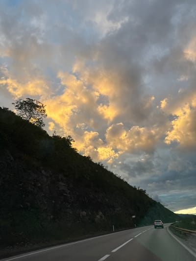 A tilted landscape view reveals a road winding alongside a mountainside under a cloudy sky in Zagorak, Montenegro. A vehicle is traveling on the road. The clouds are illuminated with gold hues. The mountainside is covered with dense vegetation and trees.