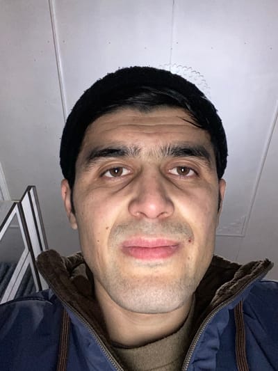 A man faces the camera wearing a dark beanie and a blue jacket with a brown collar. His eyes are brown. He has short dark hair visible under the beanie. The lighting is dim. A white ceiling and a glimpse of furniture are visible in the background. The photo has been rotated.