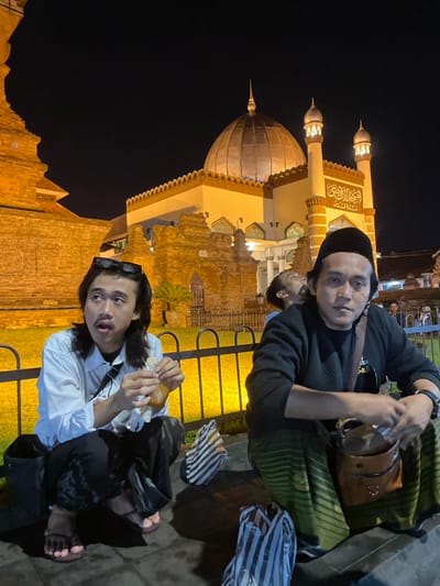This night image depicts three men in Kauman, Indonesia, with the brightly illuminated Kauman Great Mosque as a prominent background element. Two men are squatting on dark pavement in the immediate foreground, positioned behind a black metal railing that separates them from a grassy area.

The man on the left, with dark, medium-length hair, a mustache, and dark sunglasses resting on his head, wears a white long-sleeved collared shirt, dark pants, and pink flip-flops. He holds a partially consumed food item, possibly a pastry, wrapped in white paper, and looks upward and to his left with an open mouth. A black shoulder bag is slung across his body.

The man on the right, wearing a black peci (skullcap), a dark long-sleeved top (possibly a sweater or cardigan), and a green striped sarong, has a brown leather shoulder bag slung over his right shoulder and a watch on his left wrist. He looks directly forward with a neutral expression.

A third male figure is partially visible in the mid-ground, positioned between the two foreground subjects. His head is tilted sharply backward, looking upward with an open mouth, wearing a lighter-colored shirt.

The Kauman Great Mosque (Masjid Gedhe Kauman) dominates the background. It features a large, dark dome topped with a finial and two ornate minarets on its right side, both brightly lit. The mosque's facade is illuminated, revealing white walls, intricate details, and Arabic calligraphy. Older, weathered brick structures are visible to the left and center-left of the mosque, adjacent to its main building. The grassy area beyond the railing is also lit, with a small palm-like plant visible. Several striped plastic bags are on the pavement near the squatting men in the foreground.