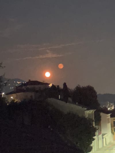 The image captures a nighttime scene in Balerna, Switzerland, with a dark sky and a faint haze. Two celestial bodies, appearing as full moons, are visible in the sky. One moon, a bright orange orb, is positioned higher and to the right of the other. The second moon, slightly dimmer and with a reddish hue, is located below and to the left of the first.

In the foreground, silhouetted against the dimly lit sky, are the rooftops of buildings and a dense cluster of trees. The architecture suggests a European urban or semi-urban setting. Distant lights from what appear to be streetlights or windows twinkle on a hillside in the mid-ground, indicating the presence of a town or settlement. The overall atmosphere is serene and slightly ethereal due to the prominent lunar presence. There are no people or discernible animals visible in the frame.