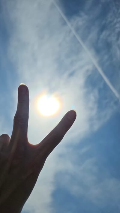 A hand with two fingers extended in a V-shape is silhouetted against a bright sun in a blue sky with scattered clouds. A faint contrail from an aircraft is visible diagonally across the sky. The image was captured at an upward angle, implying the photographer was looking up at the sky. The location is specified as Tash-Asty, Russia.