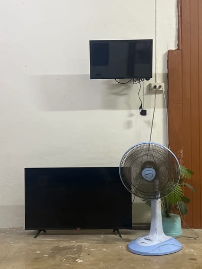 The image depicts an indoor scene, possibly a room in Thung Tako, Thailand. A white wall serves as the backdrop for two black flat-screen televisions. One television is mounted high on the wall, while the other rests on the floor. A blue and white electric fan, branded "SHARP," stands to the right of the lower television. The fan has a circular grille and a stand. Behind the fan, a green potted plant is visible, with its leaves extending upwards. Electrical outlets and cords are present on the wall near the upper television, with a power adapter hanging below. The floor appears to be concrete with some scattered white debris. A section of a brown wooden doorframe is visible on the far right edge of the image.