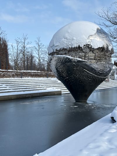 The image depicts a winter scene in the city of Plodorodny, Russia, on a clear day under a light blue sky. A large, metallic, spherical sculpture, textured and reflective, serves as the central subject. The upper portion of the sculpture is heavily blanketed with white snow, showing uneven edges and formations suggestive of melting or refreezing. Its lower half is dark and textured, tapering downwards into a conical base. This sculpture is situated within a body of water, which is predominantly frozen with a smooth, dark sheet of ice. Immediately surrounding the sculpture's base, the ice appears slightly disturbed or melted.
To the left and rear of the sculpture, a series of wide, shallow, snow-covered stone or concrete steps ascend from the water's edge. Beyond these steps, a landscaped area is visible, featuring numerous bare deciduous trees and some darker, possibly evergreen, taller trees or shrubs. A low, snow-covered hedge or embankment runs horizontally along the base of this tree line. Several dark, slender vertical posts are spaced throughout the background landscaping. In the immediate foreground, to the right, a bank of deep white snow slopes diagonally upwards from the frozen water. No individuals are present in the scene, and no overt actions or interactions are depicted.