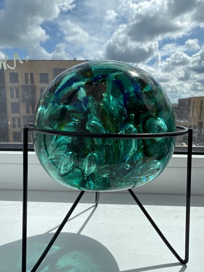 A teal glass orb filled with teal and dark blue glass shards sits on a black metal stand. The orb is positioned on a white windowsill, with a window behind it showing a sky filled with white, fluffy clouds and a hint of blue. Beyond the window, buildings are visible, with one appearing to be under construction and another with a lighter facade. The scene is illuminated by natural light, casting shadows from the stand onto the windowsill. The overall setting appears to be an indoor location overlooking a city environment.