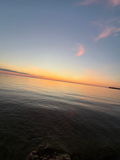 The image depicts a wide expanse of water, likely a lake or river, at sunset. The sky transitions from a deep blue at the top to soft orange and yellow hues near the horizon. A few wispy pink clouds are scattered across the upper portion of the sky. The water's surface is calm, with gentle ripples reflecting the colors of the sunset. In the foreground, a dark, rocky outcrop is visible, partially submerged in the water. The overall impression is one of tranquility and natural beauty. The location is described as being in the city of Vyshhorod, Ukraine.