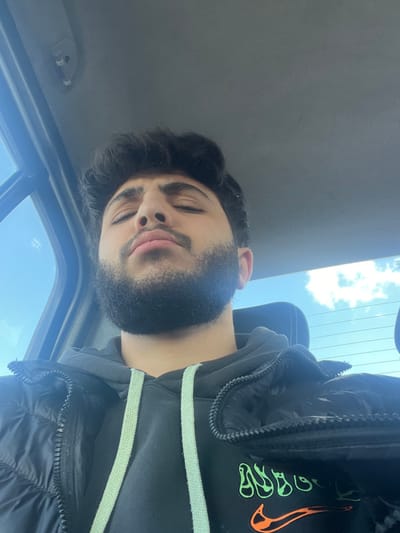 A male individual is depicted in a close-up, chest-up view, seated inside what appears to be a vehicle in the city of Al-Kiswa, Syria. He has dark, wavy hair, a dark full beard, and a mustache. His eyes are closed, and his head is tilted slightly upwards. He is wearing a black hooded sweatshirt with light green drawstrings, over which a black puffer jacket is worn, partially unzipped. A partial orange Nike swoosh logo and green abstract graphics are visible on the front of the sweatshirt. The vehicle's light-colored ceiling liner is visible at the top of the frame. Through the vehicle's windows, a clear blue sky with some scattered white clouds is visible, indicating daytime. The individual appears to be resting or sleeping.