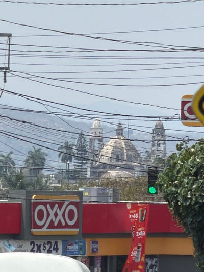 The image, taken in Cuautla, Mexico, shows a partially obscured view of the city's landmarks. A church dome and tower can be seen through a dense network of overhead electrical wires. A green traffic light is illuminated near a tree canopy. In the foreground, the side of an OXXO convenience store is visible, with advertisements for drinks and snacks. The OXXO's signage is prominent, featuring the brand's logo in red and gold, with the store's exterior colored in red, yellow, and gray. The overall image captures the blend of urban infrastructure, commercial elements, and historical architecture in Cuautla.