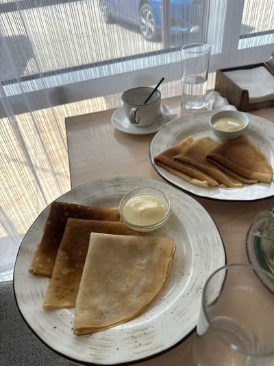 The image displays two plates of crepes with bowls of custard on a wooden table, set against a backdrop of sheer white curtains. The curtains are illuminated by sunlight, casting striped shadows on the table and the floor, and a faint reflection of a blue car is visible through them. In the background, a glass of water, a cup and saucer with a straw, and a napkin holder are also present. The scene suggests a meal or snack being enjoyed indoors, possibly at a cafe or restaurant. The location is identified as Slyudyanka, Russia.