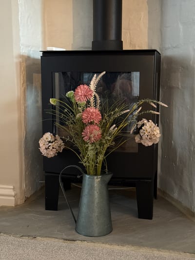 The image displays an indoor scene featuring a black wood-burning stove, positioned in what appears to be a fireplace alcove or corner of a room in Harrogate, United Kingdom. The stove is square-shaped with a glass door in the front, reflecting a subtle, indistinct interior. A black stovepipe extends from its left side.

On the right side of the stove, a galvanized metal pitcher or jug, with a handle, holds an arrangement of artificial flowers. The bouquet includes prominent pink, multi-petal blossoms, light cream or white hydrangeas or similar clusters, and tall, wispy green foliage, possibly lavender or similar dried plants, adding texture and height. Some of the flowers and foliage spill over the front and top of the stove.

The surrounding walls are textured, appearing to be painted brick or rough-rendered plaster. The wall to the left of the stove is a warm, light cream, while the upper section and the wall to the right exhibit a lighter, off-white or light grey hue. The floor surface in the immediate foreground and under the stove is a light-colored, possibly stone or concrete material, transitioning to a light grey carpet or rug on the right side of the image. The lighting is soft and ambient, suggesting an indoor setting without direct sunlight, and no specific time of day is discernible. There are no people visible, no activity taking place, and no discernible text. The overall scene suggests a cozy, decorative, and rustic interior setting.